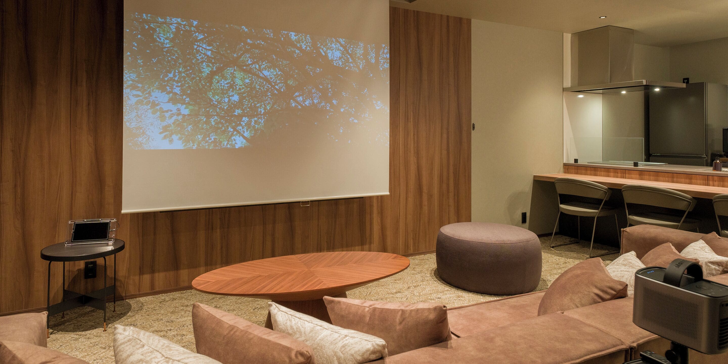 A modern conference room with a large projection screen, a round wooden table, and comfortable seating.