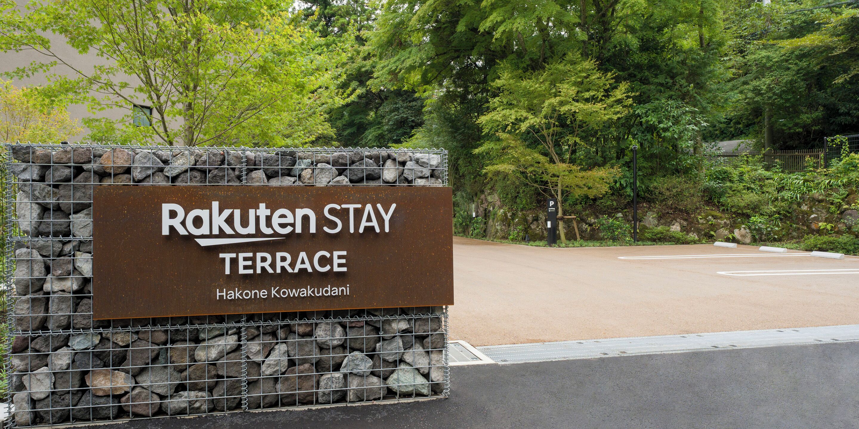 A sign for Rakuten Stay Terrace with a stone wall and a parking area in the background.