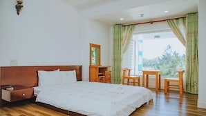 A bedroom with a wooden bed, a nightstand, a desk, and a chair. There is a window with curtains and a view of trees outside.