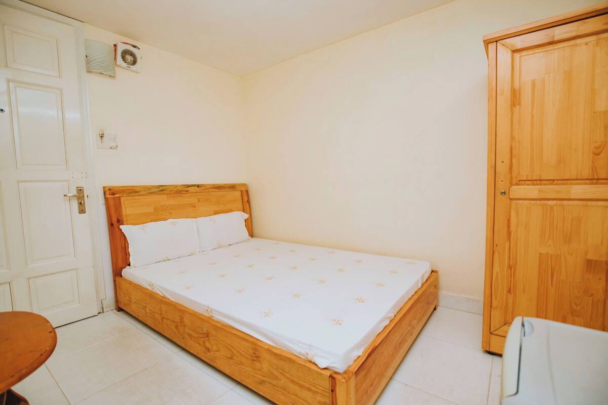A single-bed room with a wooden headboard, a white bedspread, a wooden wardrobe, and a small table.
