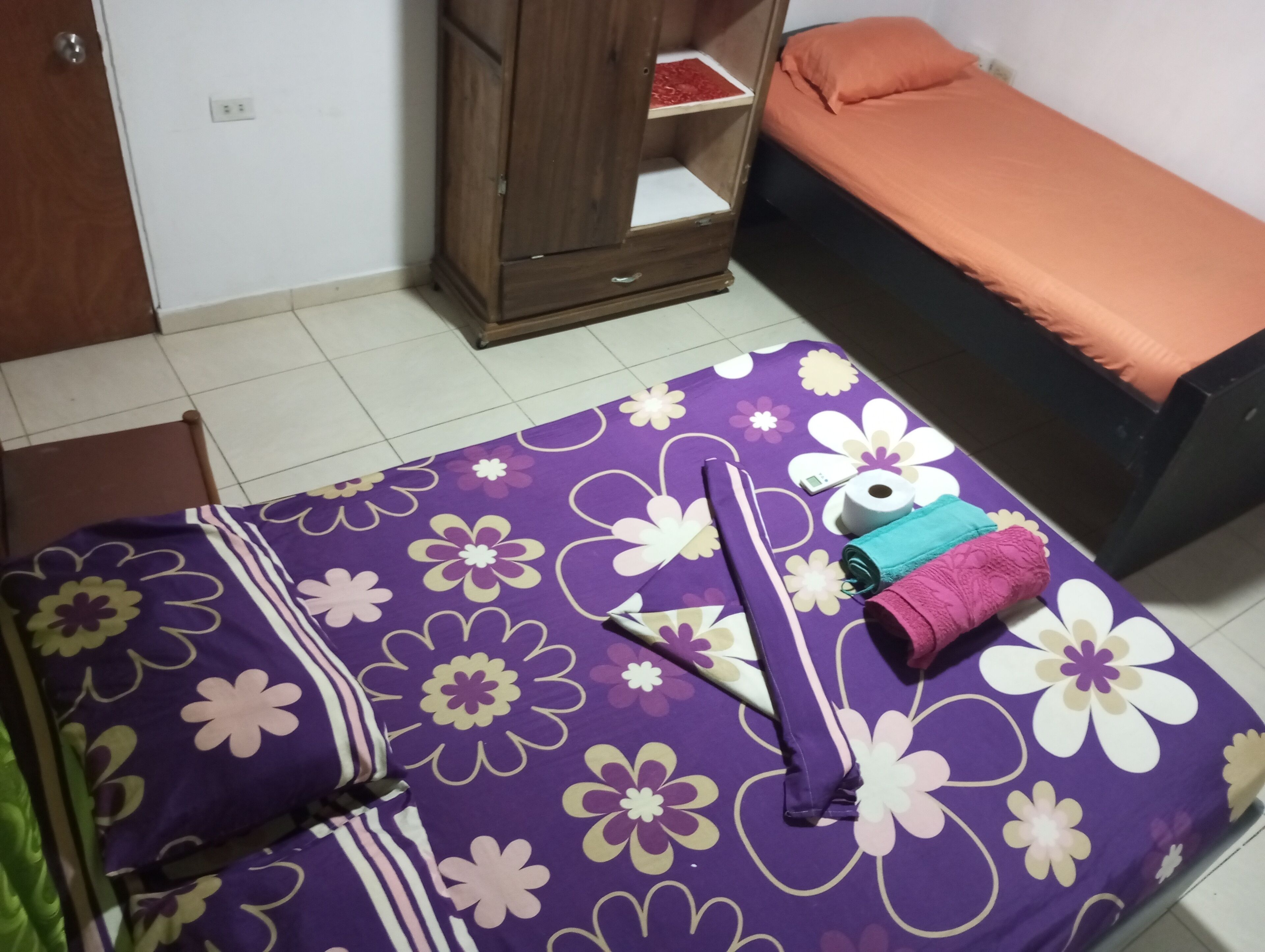 A room with a bed covered in a purple floral bedspread, a wooden wardrobe, and a small orange sofa.