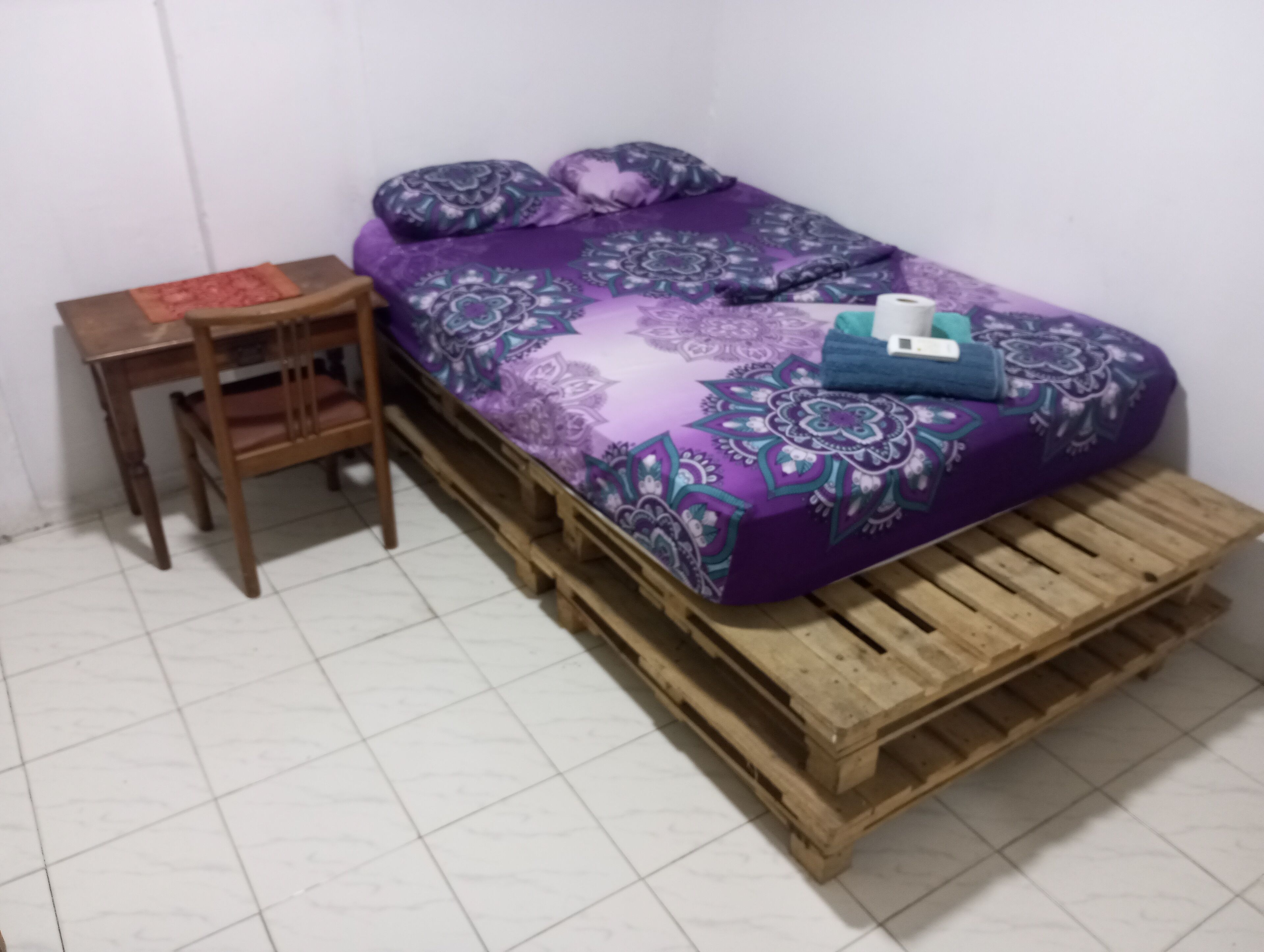Een kleine kamer met een bed op een houten pallet, een houten stoel en een tafeltje met een rood voorwerp erop.