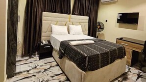 A hotel room with a large bed, a desk, a chair, a TV, and an air conditioning unit.