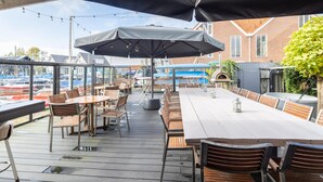 An outdoor patio with wooden tables and chairs, umbrellas, and a view of boats and buildings in the background.