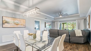 A modern living room with a glass dining table, white chairs, a gray sectional sofa, and a ceiling fan. - Villas South Meadow: Free Water Park, Golf View, Pool, Near Disney (Kissimmee)