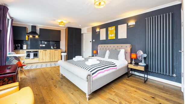 A modern hotel room with a large bed, a bedside table with a lamp, a small table with a chair, and a view of the kitchen and living area.