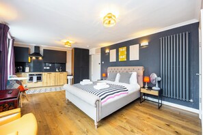 Una habitación de hotel moderna con una cama grande, una mesita de noche con una lámpara, una pequeña mesa con una silla y vistas a la cocina y la sala de estar.