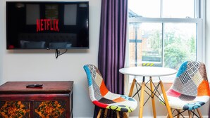 A room with a TV displaying 'NETFLIX', a wooden table, two colorful chairs, and a window with a view of buildings and trees.