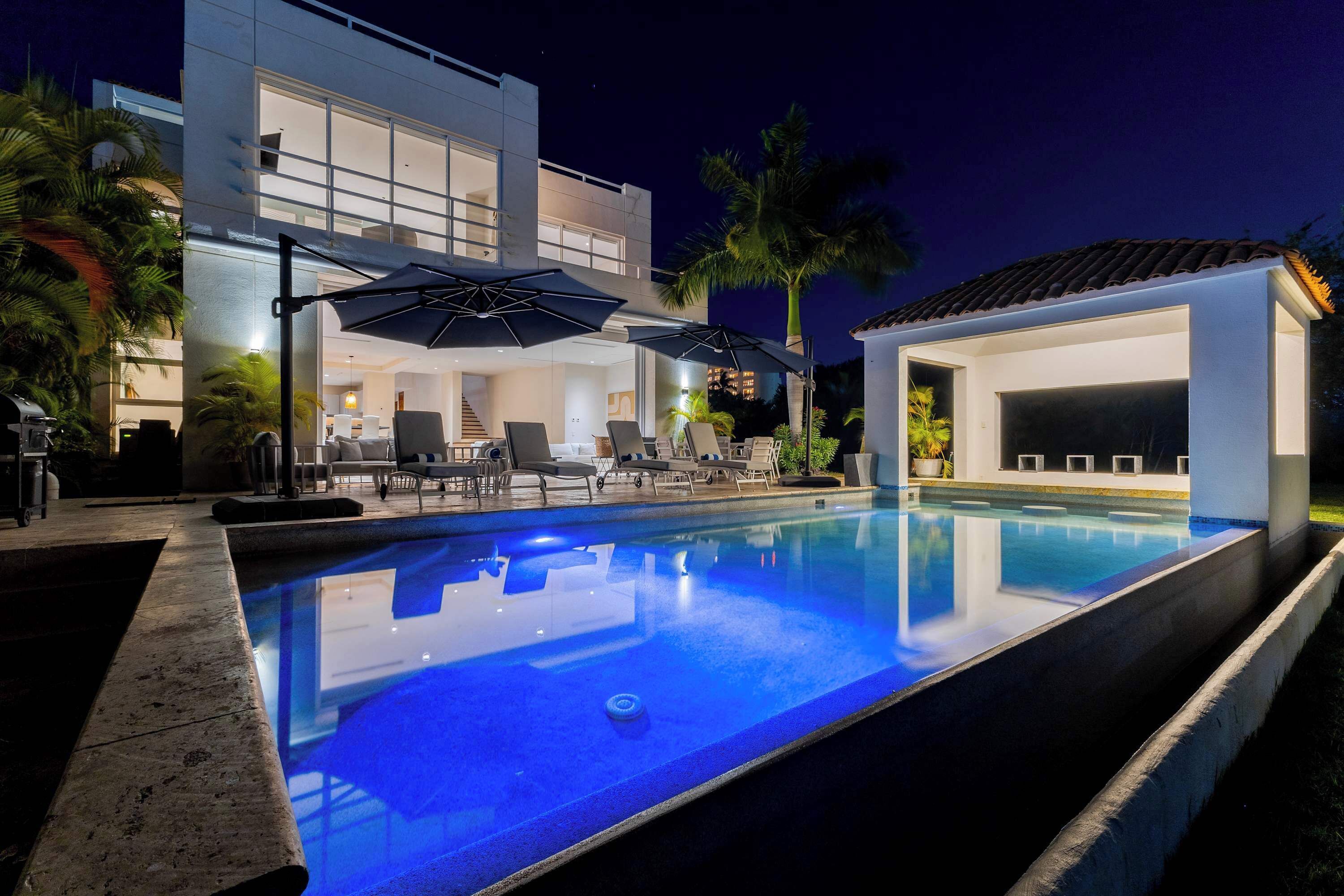 Un espace piscine moderne avec des chaises longues et des parasols, une piscine et un bâtiment doté de grandes fenêtres et d’un balcon.