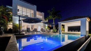 Et moderne poolområde med liggestole og parasoller, en pool samt en bygning med store vinduer og en balkon.