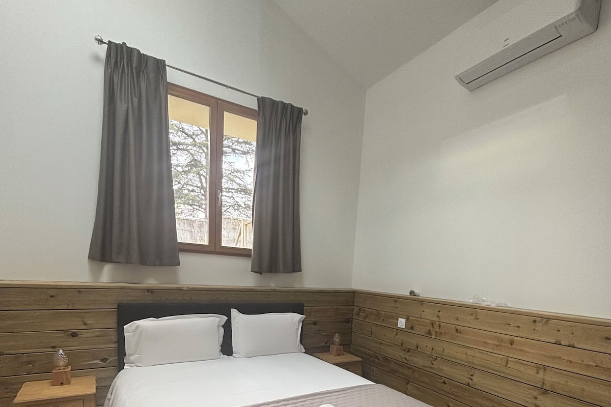 A bedroom with a wooden headboard, a bed with white linens, a nightstand with a lamp, and a window with curtains.