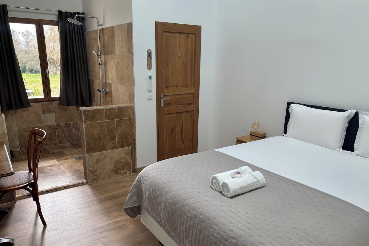 A bedroom with a bed, a wooden door, a shower area, and a chair.