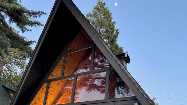 A modern, triangular wooden cabin with large glass windows reflecting the surrounding trees and sky. - A-Frame with Carousel Fireplace - escape to the mountains and get out in natu... (Running  Springs)