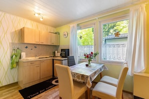 A compact kitchen with wooden cabinets, a dining table with chairs, a small window with a view of greenery, and a clock on the wall.