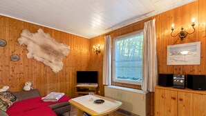 Habitació acollidora amb parets de fusta, televisió, taula amb mantell vermell i finestra amb cortines.