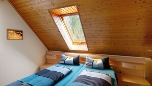 A room with a sloped ceiling, a skylight, two beds with blue bedding, and a small bedside table.