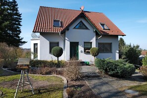 Une maison à deux étages, avec un toit recouvert de tuiles rouges, un petit balcon et un jardin où les arbustes sont taillés et où se trouve un banc en bois.