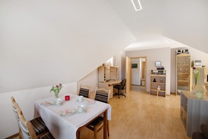 A dining area with a table set for two, a small plant, and a red cup. There is a desk with a chair and a bookshelf in the background.