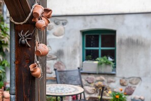 Ein rustikales Freigelände mit einem Holzpfosten, der mit Terrakottatöpfen und einer Spinnendekoration verziert ist, einer Steinsäule, einem Fenster mit grünem Rahmen und einem runden Tisch mit Stühlen.
