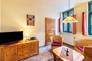 A furnished room with a television, radio, wooden cabinet, wicker chairs, a table with a bottle, and a window with curtains.
