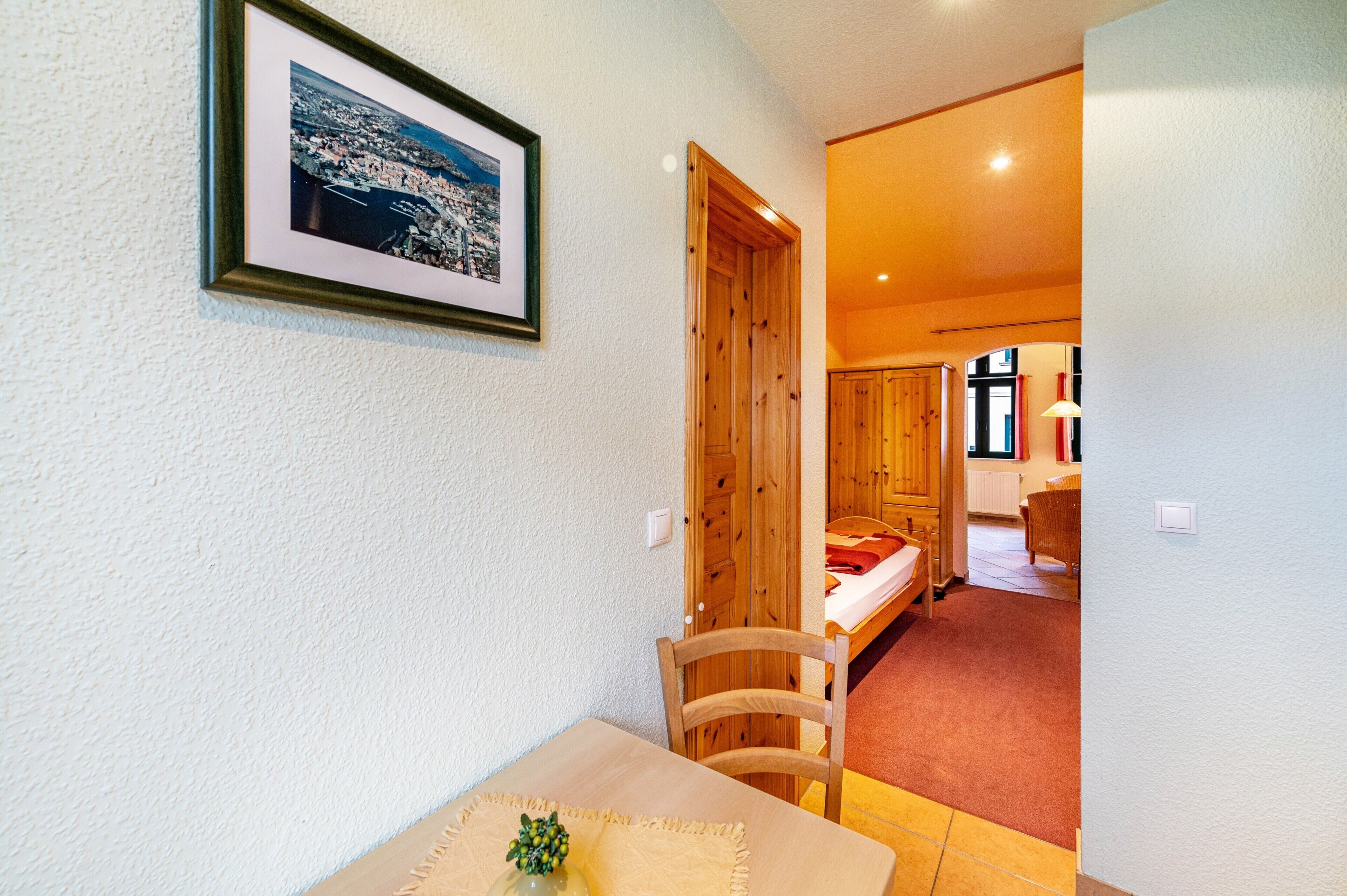 Ein Zimmer mit Holztür, einem Bett, einem kleinen Tisch mit einer Vase und einem gerahmten Bild an der Wand.