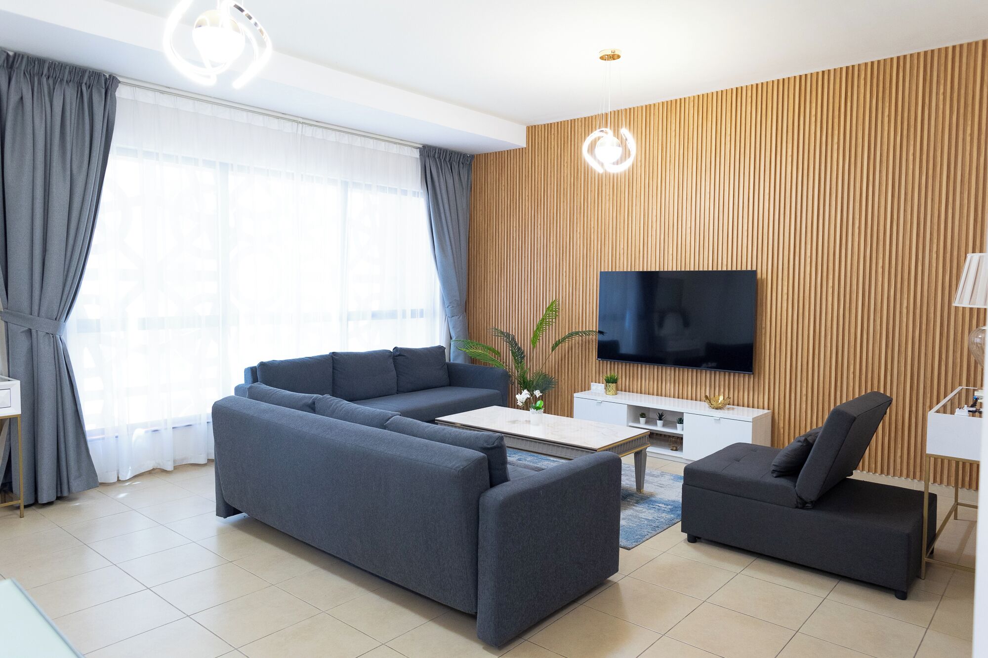 Moderni olohuone, jossa on harmaa sohva, pienempi harmaa nojatuoli, sohvapöytä, puiseen seinään kiinnitetty televisio ja valkoinen kaappi, jossa on koriste-esineitä.