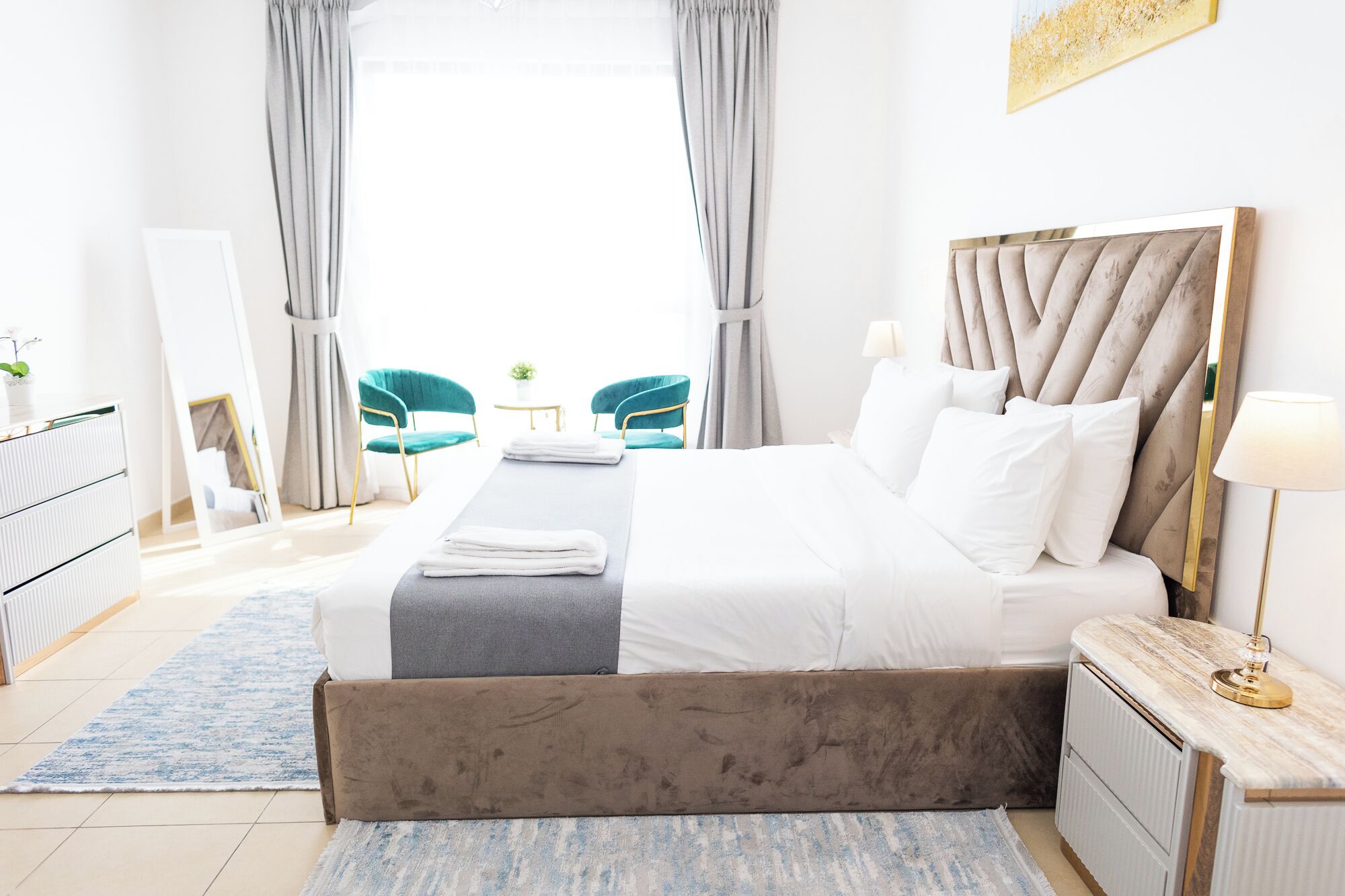 A hotel room with a bed, bedside tables, a mirror, a window with curtains, a desk, and two chairs.