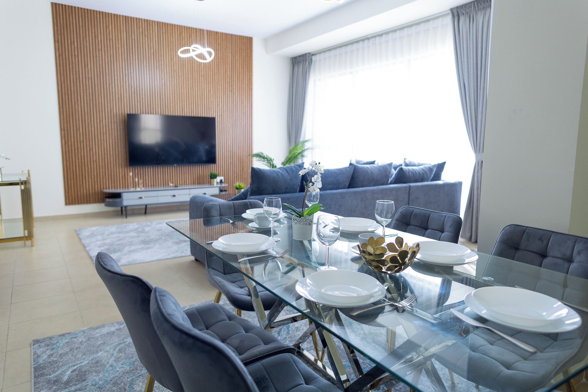 Un saló modern amb una taula de dinar de vidre preparada per a quatre persones, un sofà, un televisor muntat sobre una paret de fusta i una finestra gran amb cortines.
