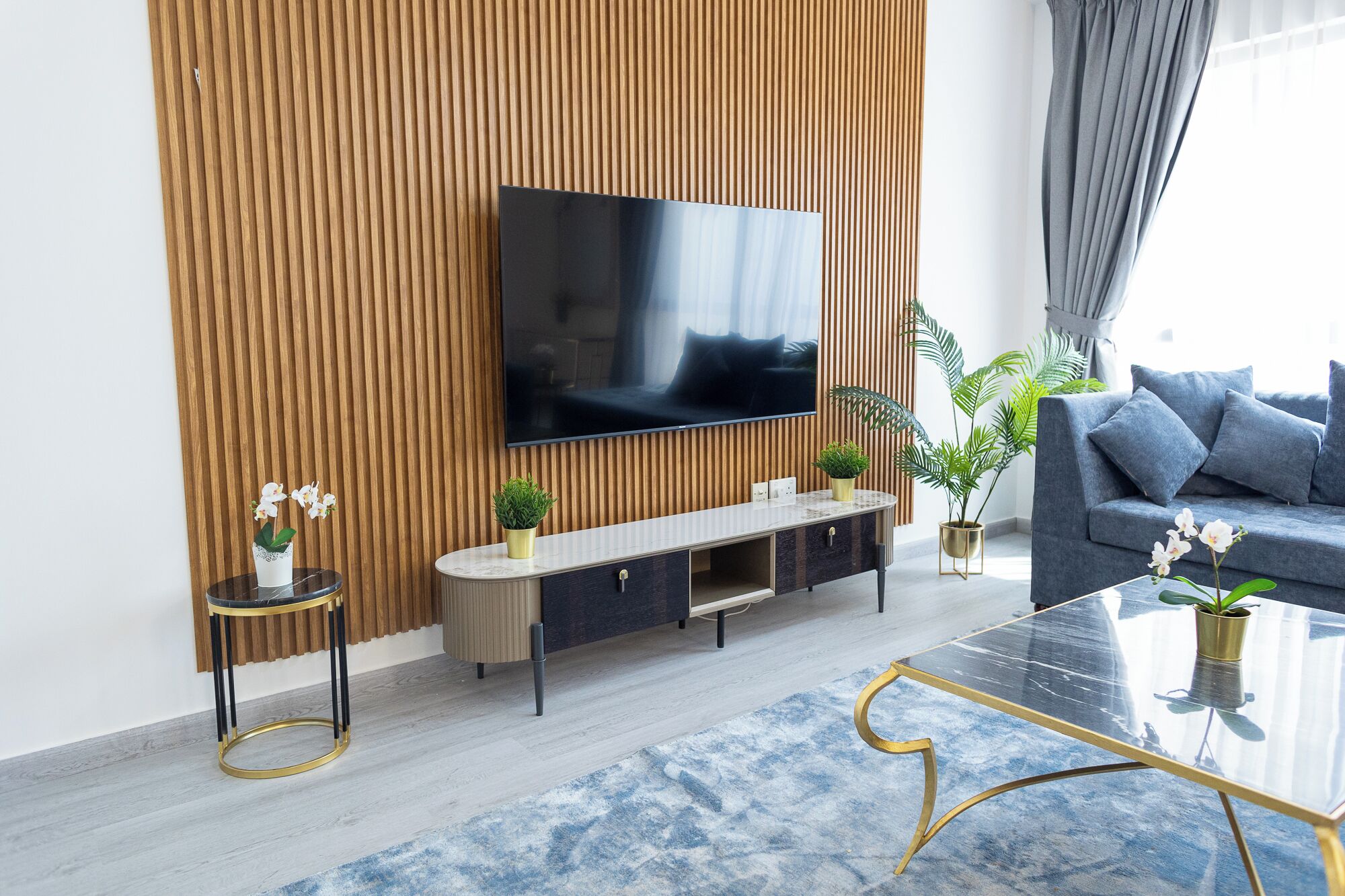 Un salon moderne avec un téléviseur à écran plat fixé sur un mur recouvert de panneaux en bois, un canapé gris, une table basse et une petite table d’appoint agrémentée d’une plante.