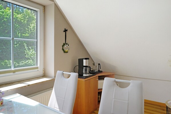 Een kleine, gezellige kamer met een tafel, twee stoelen, een koffiezetapparaat en een raam met uitzicht op bomen.