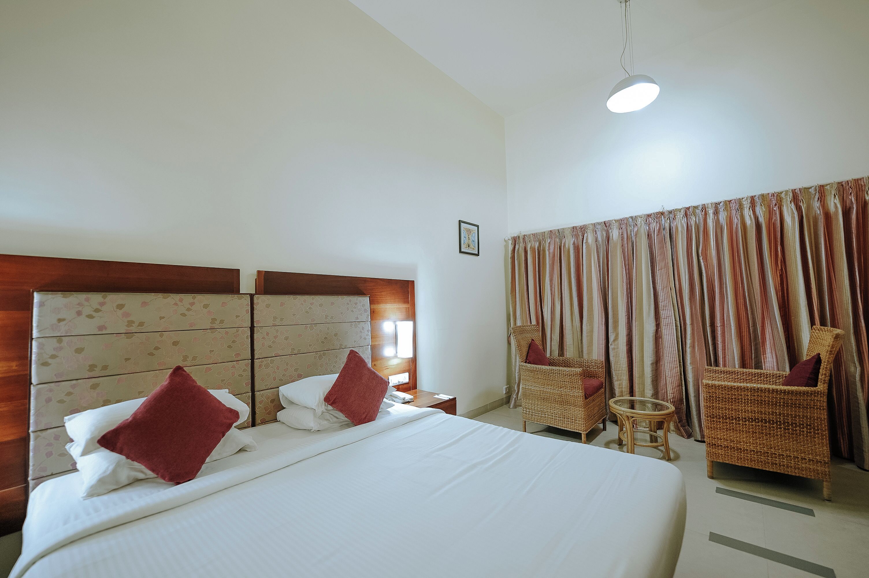A hotel room with a large bed, two nightstands, a small round table, two wicker chairs, a large window with curtains, and a ceiling light.