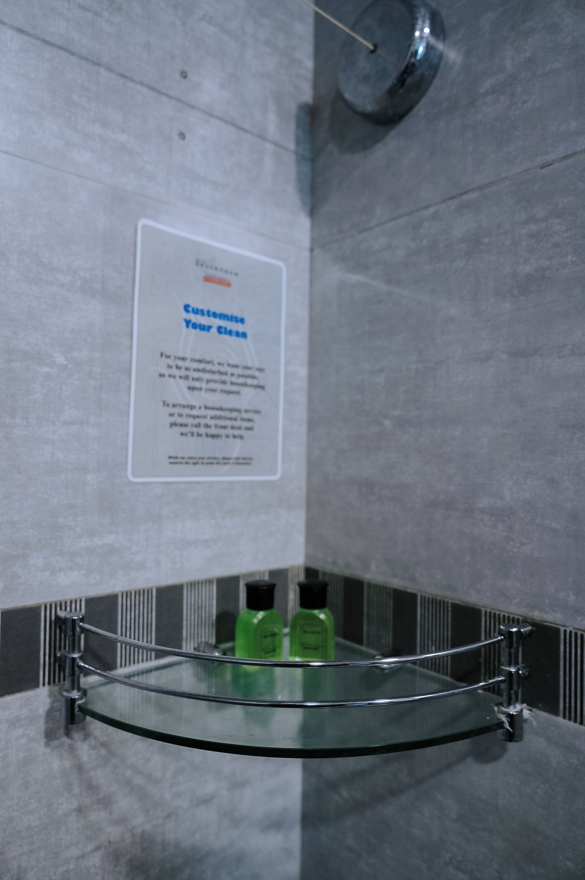 A bathroom with a glass shelf holding toiletries, a sign on the wall, and a showerhead.