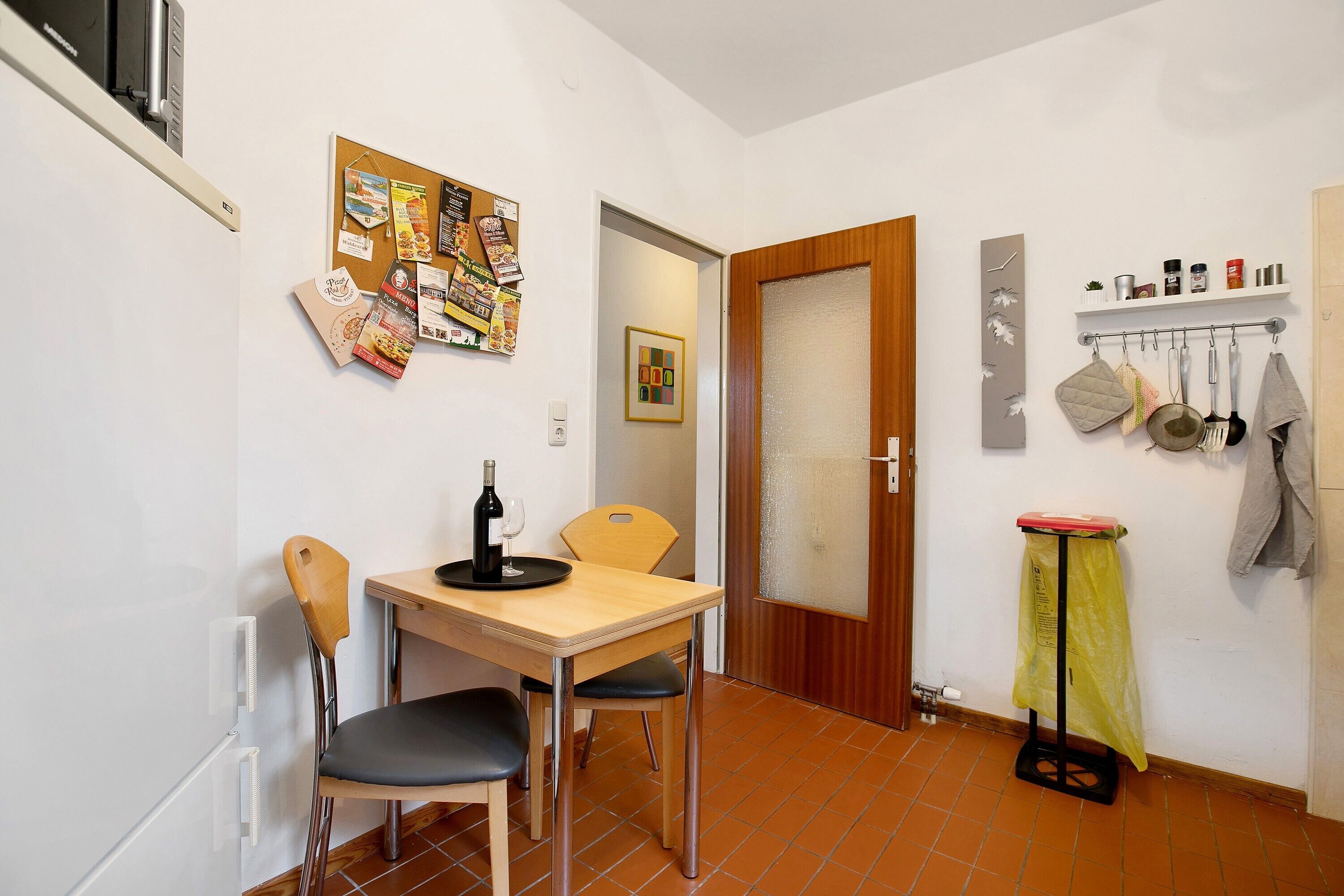 Un pequeño comedor con una mesa, sillas, una copa de vino y un tablón de anuncios con varios papeles.