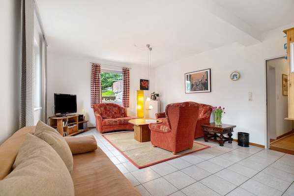 A living room with a sofa, armchair, TV stand, and a small table with a lamp.