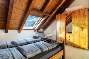 A room with a sloped ceiling, wooden beams, a skylight, a bed with patterned bedding, a small table, and a wooden wardrobe.