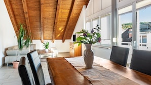 A dining area with a wooden table, chairs, a vase with flowers, and a view of the outside through large windows.