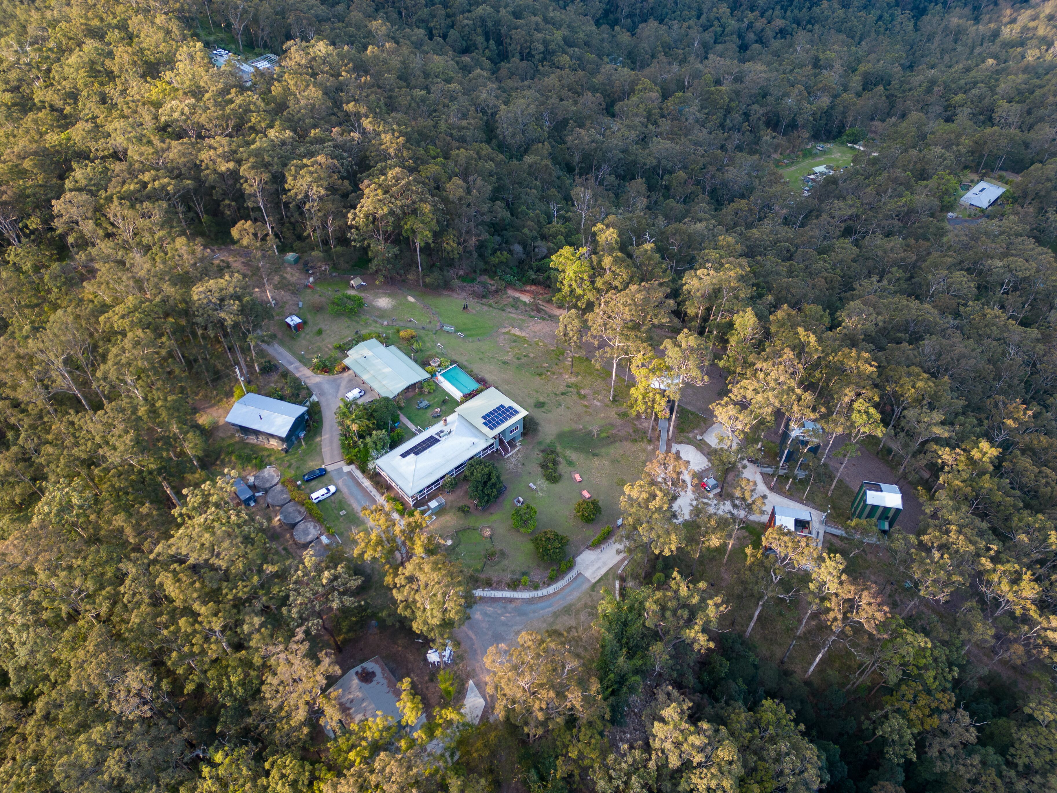 Une vue aérienne d’une propriété entourée d’une forêt dense, avec un bâtiment principal doté d’un grand toit, plusieurs constructions plus petites et une piscine bien visible.