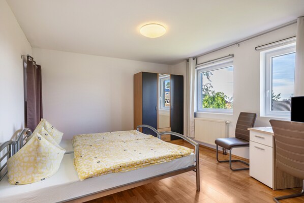 A single bed with a patterned bedspread, a desk with a chair, a radiator, and a window with curtains.