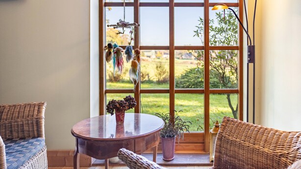 Una habitación con un ventanal grande, muebles de mimbre, una mesa redonda y una planta en maceta.