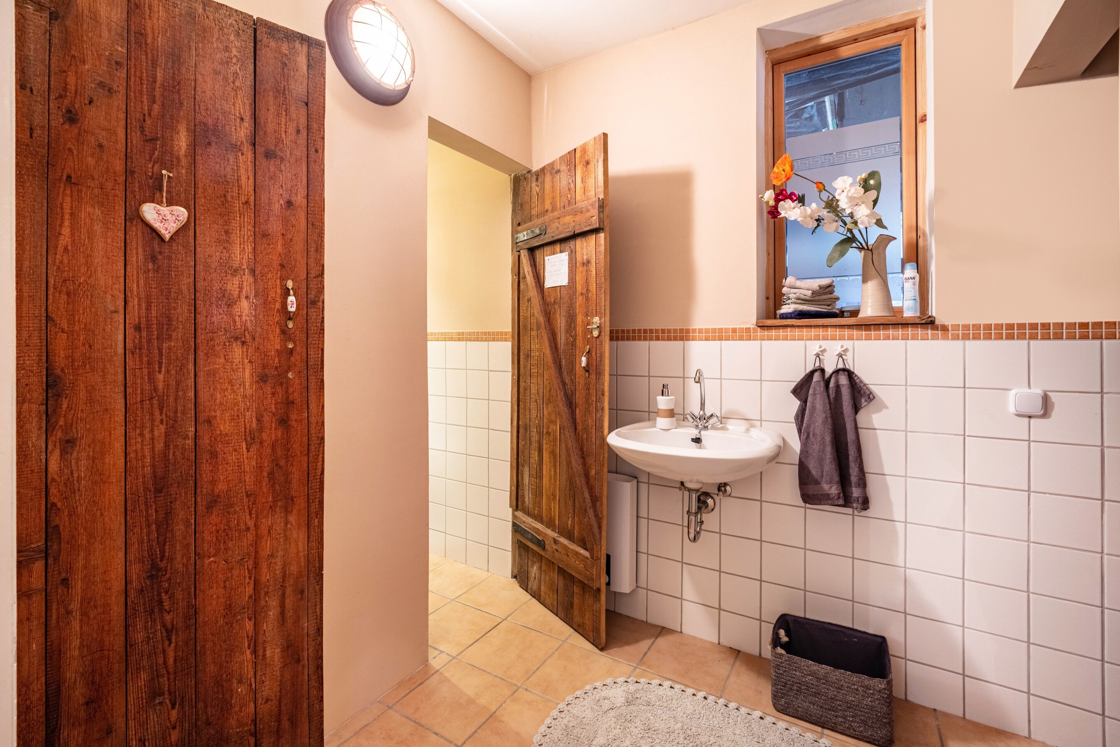 Un baño con puerta de madera, un lavamanos blanco, un espejo con un florero y un perchero para toallas.