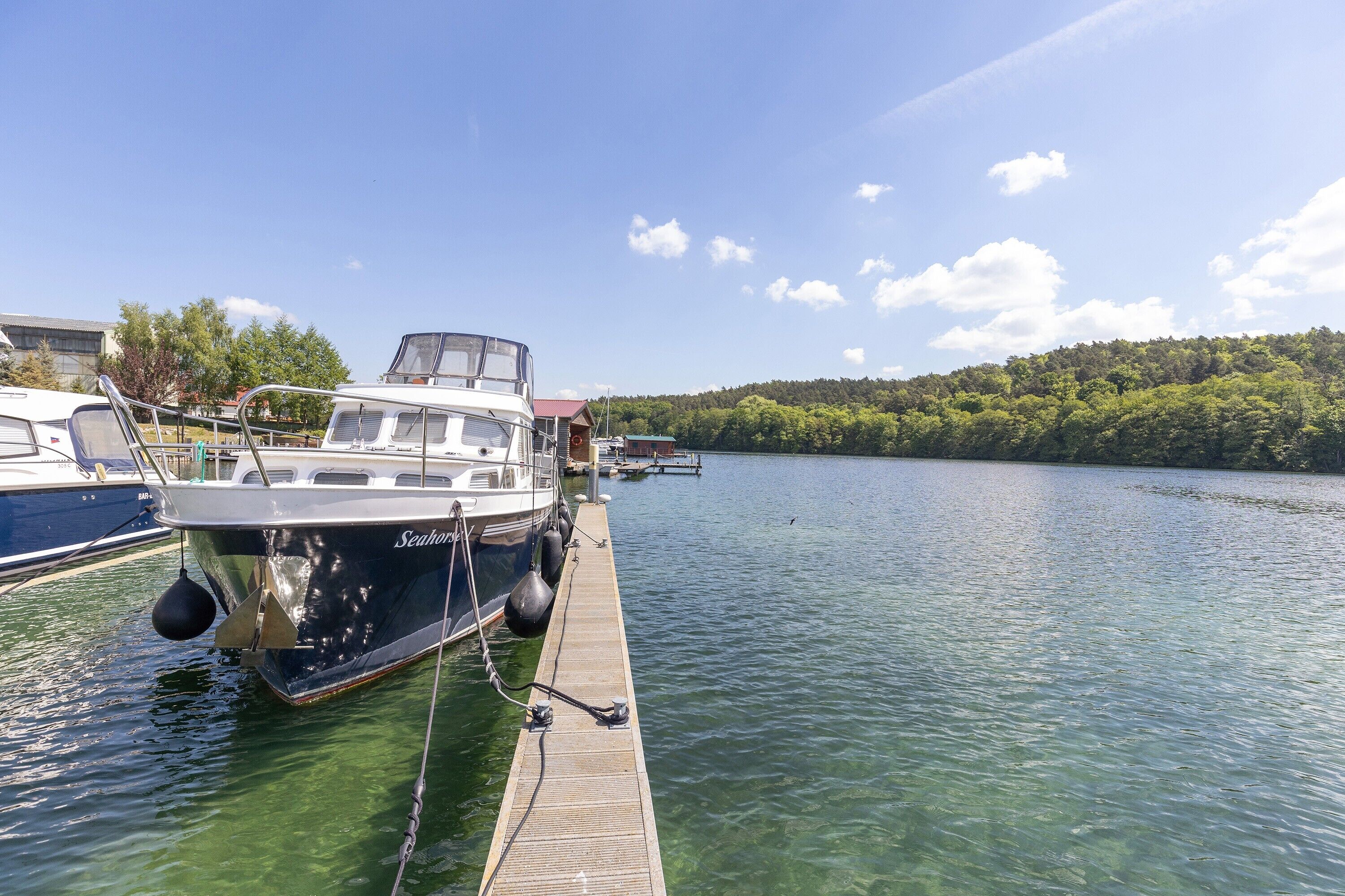A boat docked at a pier with clear water, a dock extending into the lake, and a forested hill in the background.