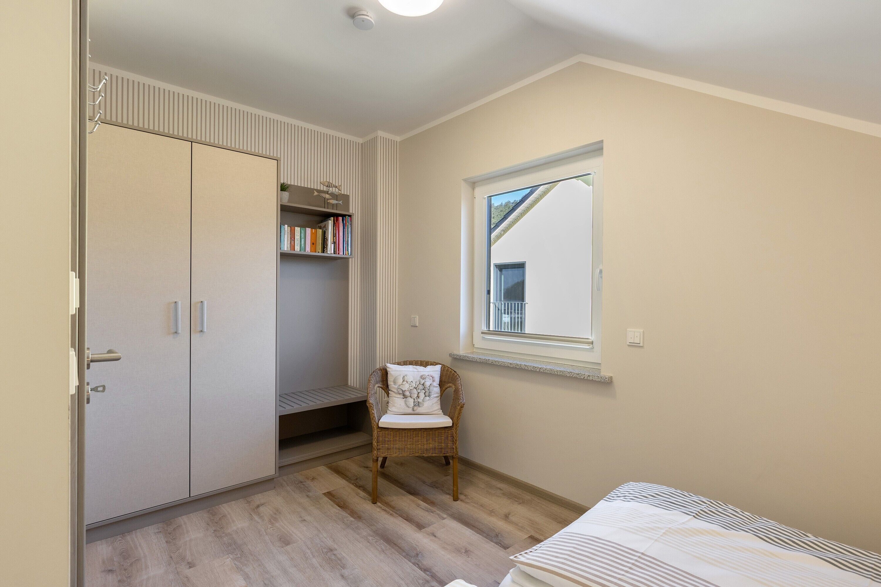 Ein Schlafzimmer mit Bett, Fenster, Sessel, Kleiderschrank und einem Regal mit Büchern.