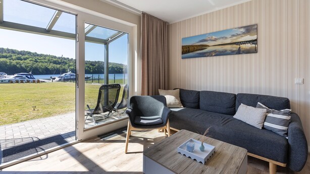 A modern living room with a sofa, armchair, and coffee table, large windows with a view of boats and greenery, and a painting on the wall. - Apartment 'Ferienwohnung 2' with Shared Garden and Wi-Fi (Joachimsthal)