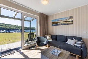 Un salon moderne avec un canapé, un fauteuil et une table basse, de grandes fenêtres offrant une vue sur les bateaux et la verdure, et un tableau accroché au mur.