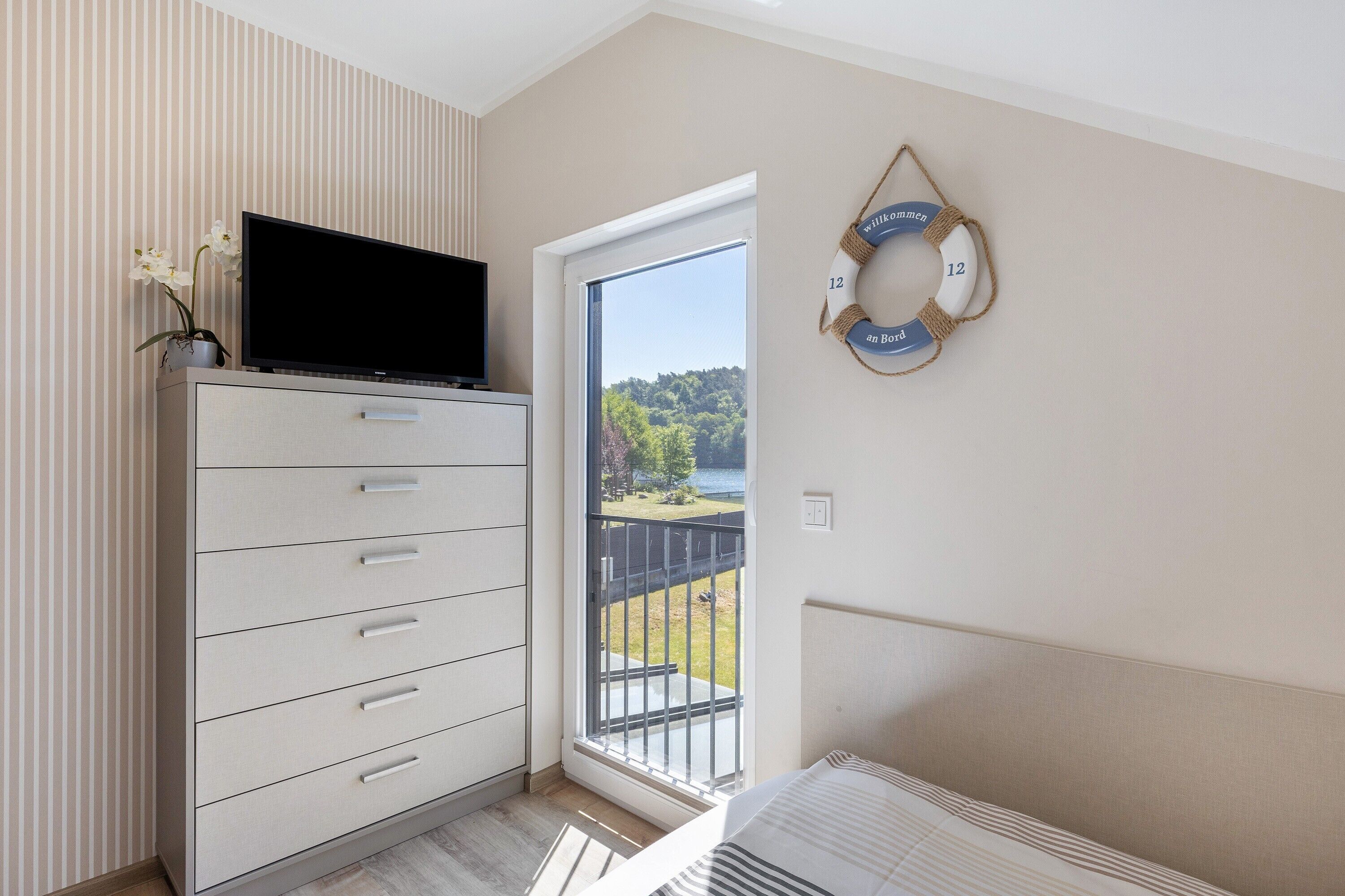 A bedroom with a TV on a dresser, a bed, a balcony with a view, and a wall clock.