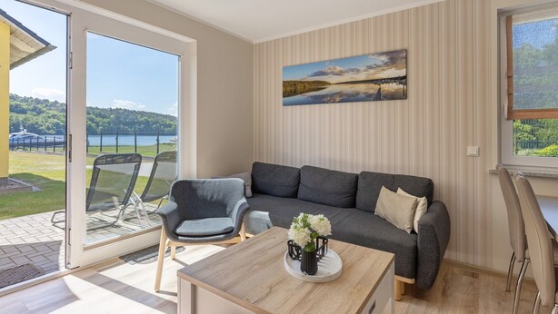 A modern living room with a grey sofa, a coffee table with a vase, and a large window with a view of a lake and hills.