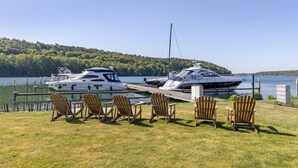 A marina with two yachts docked, a grassy area with wooden chairs, and a forested hill in the background.