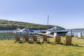 Un puerto deportivo con dos yates amarrados, una zona ajardinada con sillas de madera y una colina boscosa al fondo.
