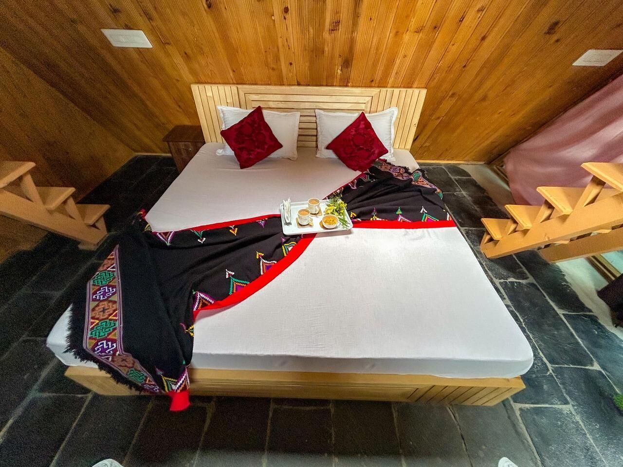 A bed with a white mattress, red pillows, and a black blanket with colorful embroidery. There is a tray with a cup and a small plate on the bed.