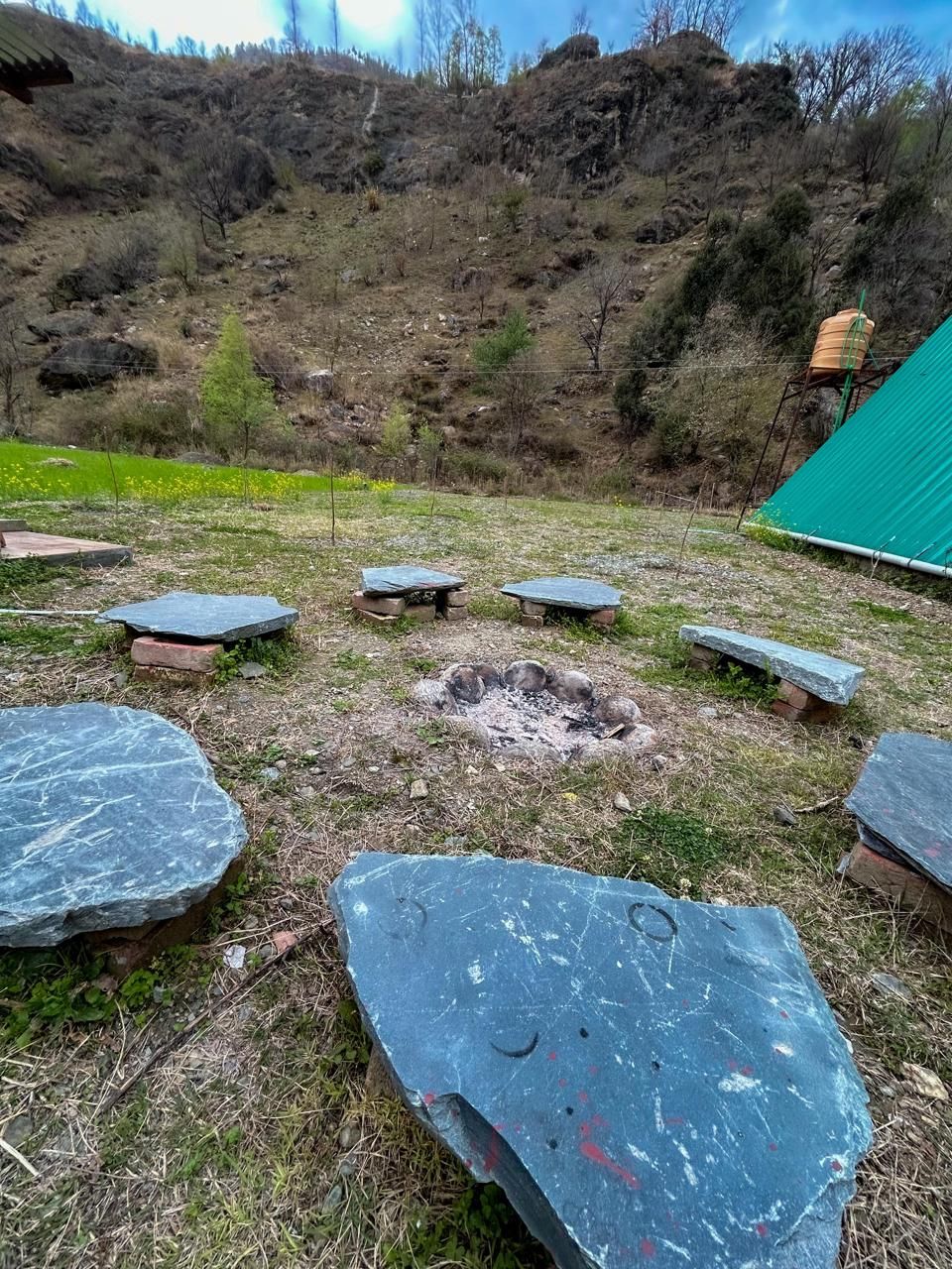Un’area relax in pietra con un braciere al centro, circondata da un prato e un pendio.